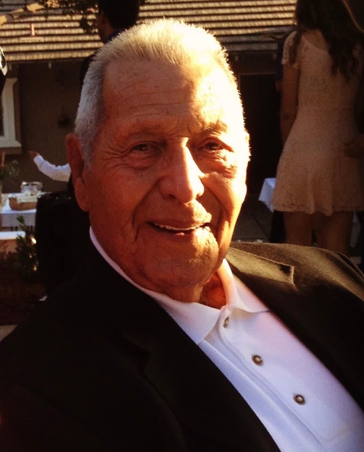 Elderly man with short grey hair smiles warmly, illuminated by golden sunlight at an outdoor gathering.